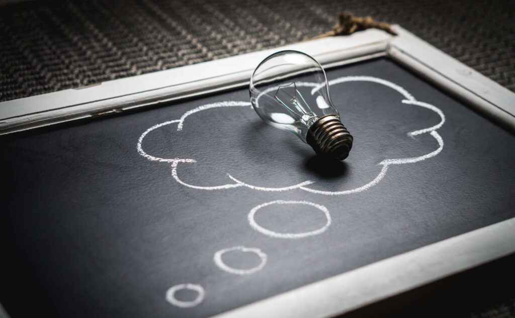 Lightbulb resting on a chalkboard with a drawing of a thought cloud.